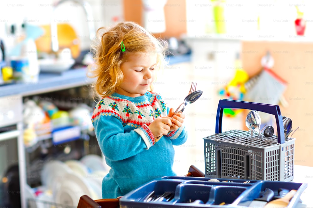 Cute little toddler girl helping in the kitchen with dish washing machine. Happy healthy blonde child sorting knives, forks, spoons, cutlery. Baby having fun with helping housework mother and father
