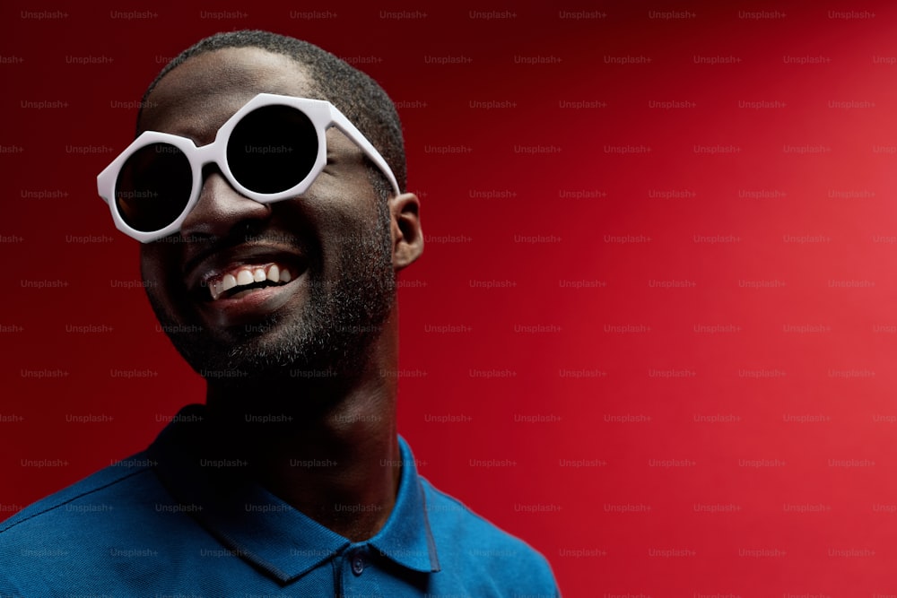 Mode. Homme noir souriant avec des lunettes de soleil sur fond rouge. Portrait contrasté d’un modèle masculin afro-américain joyeux et joyeux dans des lunettes élégantes et un sourire blanc au studio