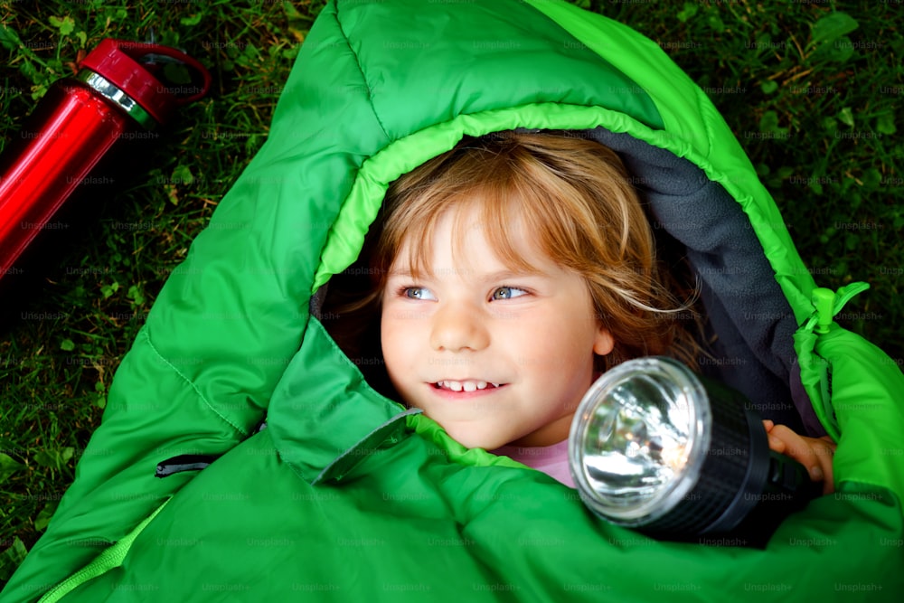 Preschool little girl in sleeping bag camping. Outdoors activity with children in summer. Fun and adventure camp, family and friends vacations or weekend trip. Portrait of child with flashlight