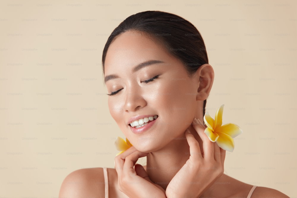 Woman And Flowers Beauty Portrait. Beautiful Ethnic Girl With Perfect Glowing And Hydrated Facial Skin. Asian Model With Closed Eyes Holding Tropical Plumeria Near Face Against Beige Background.