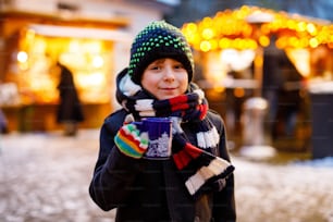 Little cute kid boy drinking hot children punch or chocolate on German Christmas market. Happy child on traditional family market in Germany, Laughing boy in colorful winter clothes.