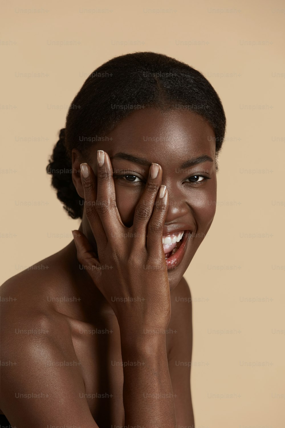 Primer plano del retrato de una hermosa chica africana. Una joven sonriente toca su rostro limpio y mira a la cámara. Concepto de cuidado de la piel del rostro. Aislado sobre fondo beige. Sesión de estudio