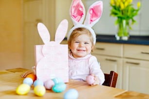 Cute little toddler girl wearing Easter bunny ears playing with colored pastel eggs. Happy baby child unpacking gifts. Adorable kid in pink clothes enjoying holiday.