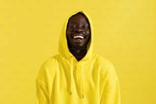 Portrait d’homme noir heureux en sweat à capuche jaune sur fond couleur. Modèle masculin afro-américain souriant et joyeux avec un sourire blanc dans un sweat-shirt élégant avec capuche sur la tête dans le studio