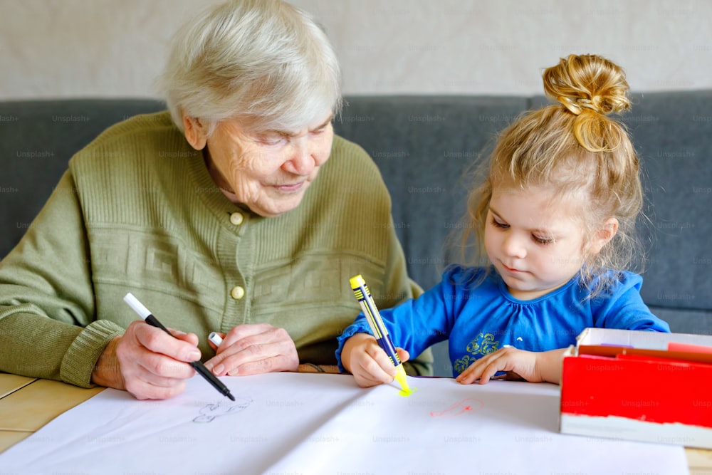 Beautiful toddler girl and grand grandmother drawing together pictures with felt pens at home. Cute child and senior woman having fun together. Happy family indoors.