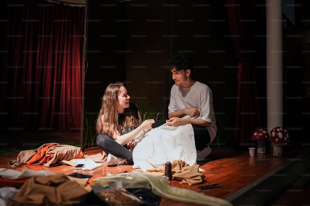 a man and a woman sitting on the floor in front of a pile of clothes