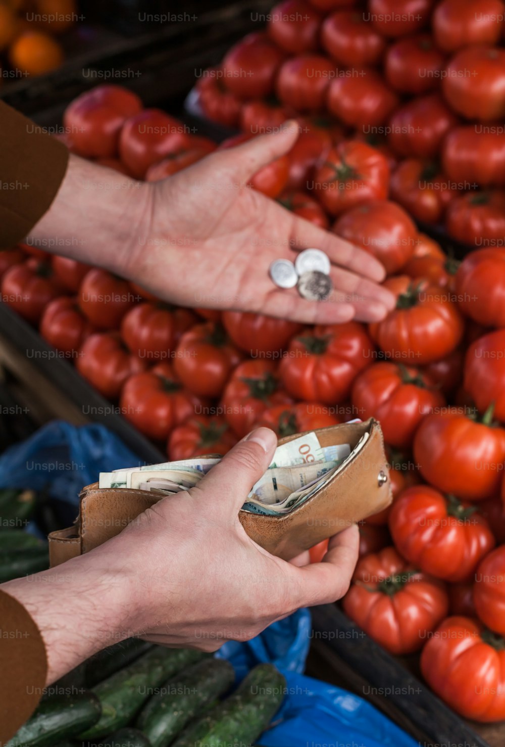 a person holding a wallet in front of a pile of tomatoes and cucumbers