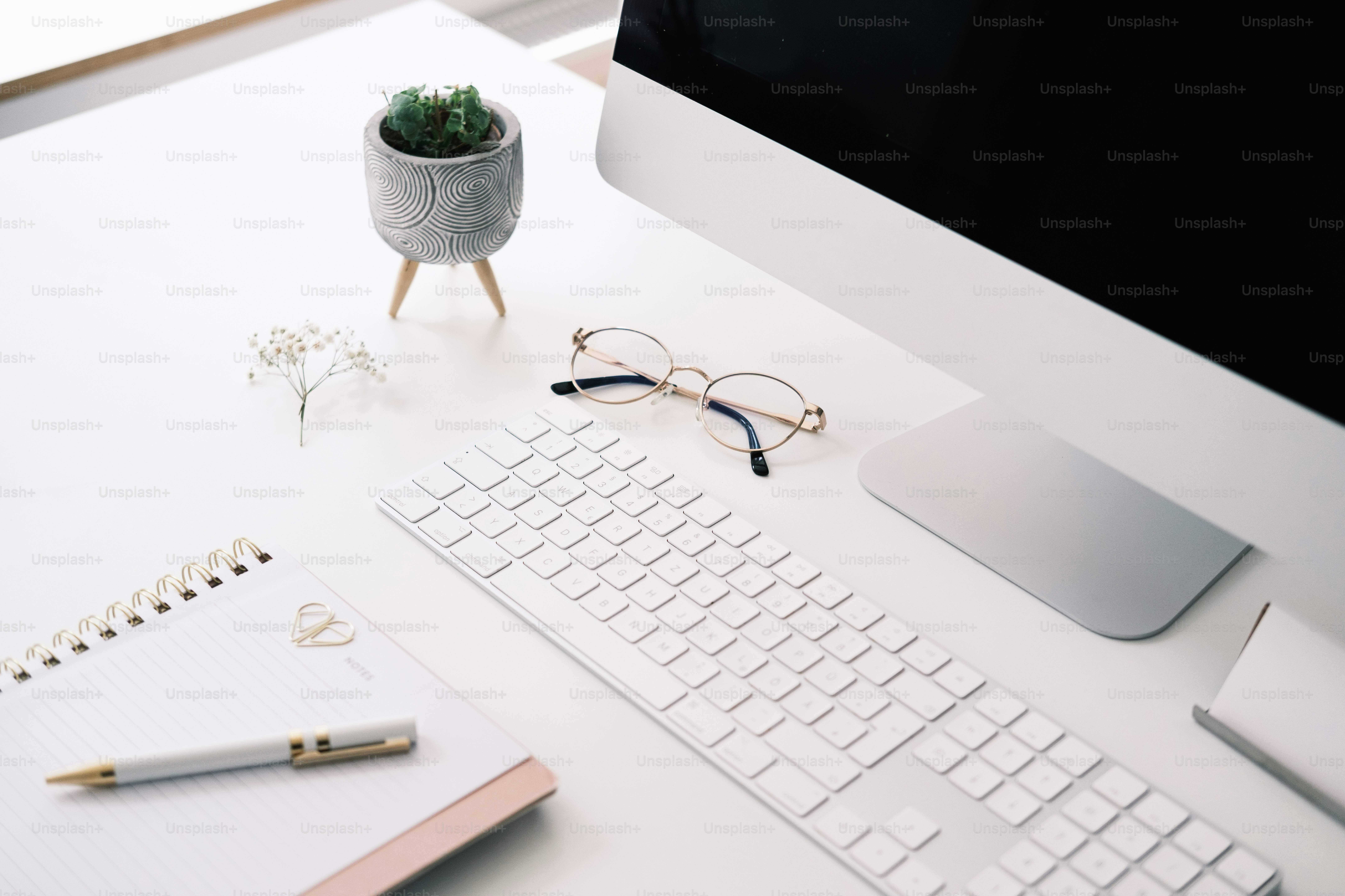 a desk with a keyboard, monitor, glasses and a plant
