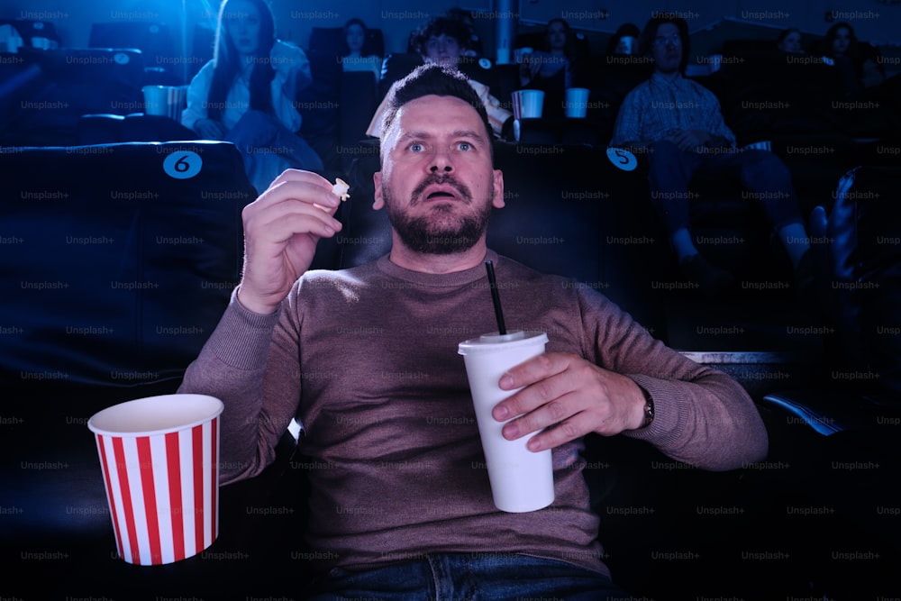 um homem sentado em um cinema segurando uma bebida e pipoca