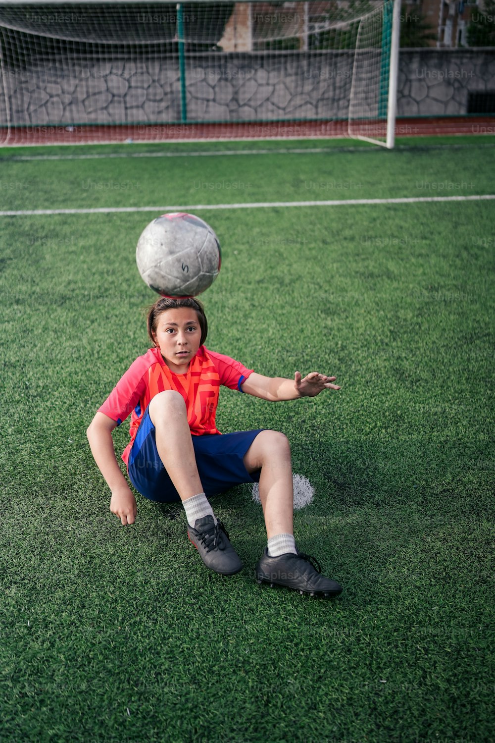 頭にサッカーボールを乗せて地面に座っている少年