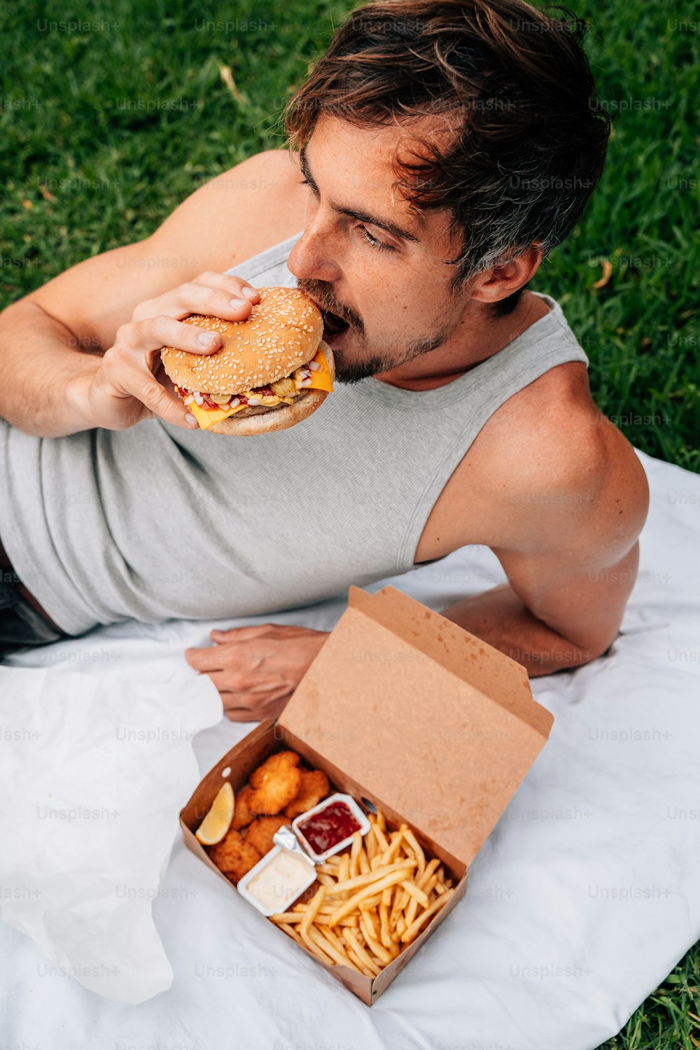 um homem deitado em um cobertor comendo um hambúrguer e batatas fritas