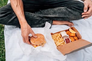 Un hombre sentado en una manta sosteniendo una hamburguesa y papas fritas