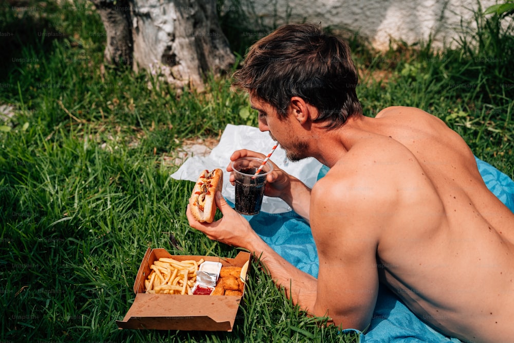 Un hombre sin camisa comiendo un perrito caliente y papas fritas
