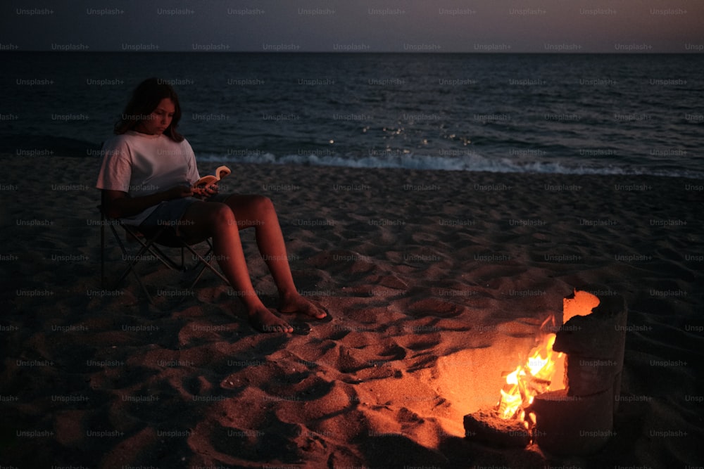 uma mulher sentada em uma cadeira em uma praia ao lado de uma fogueira