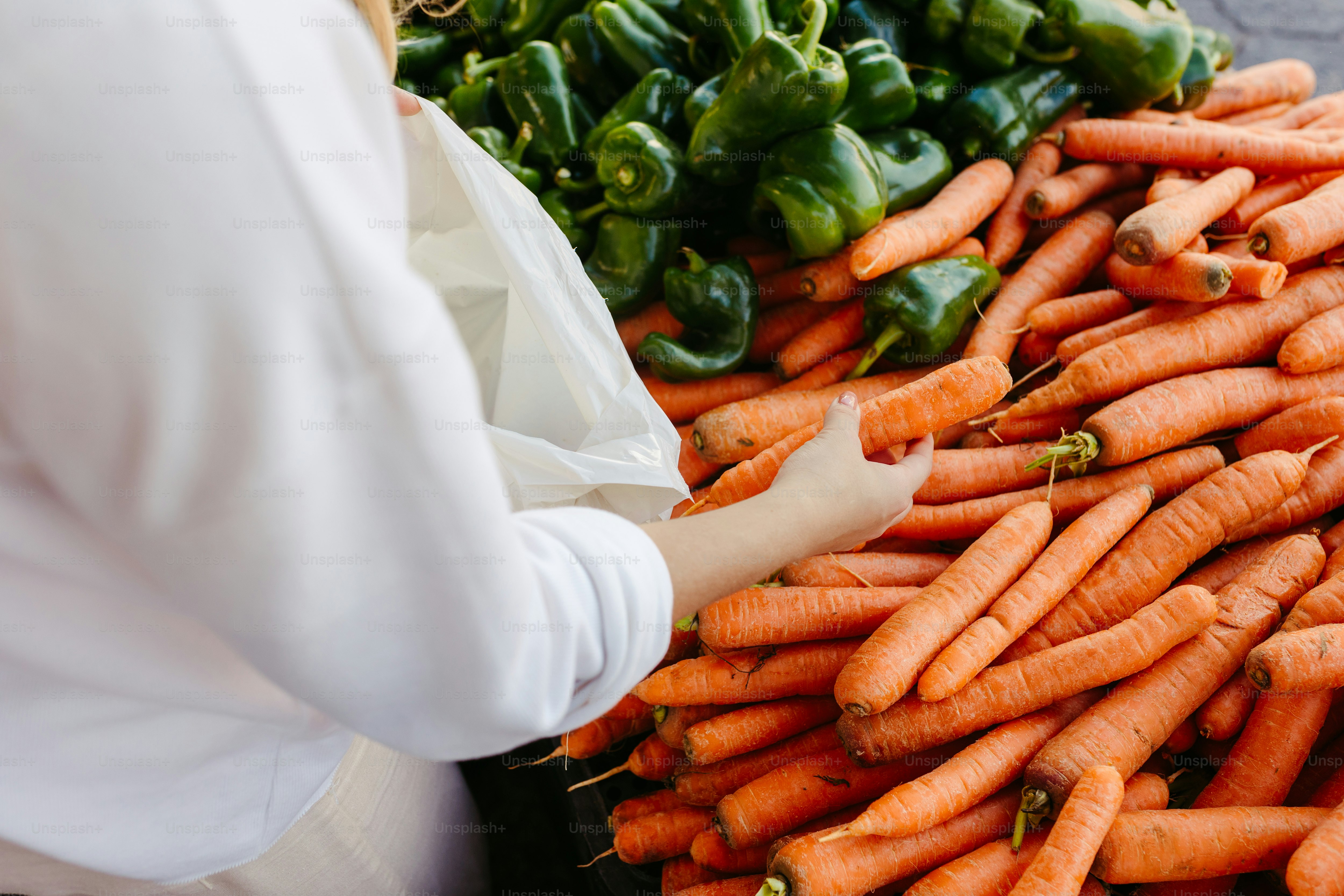 a woman holding a bag of carrots in front of a pile of peppers