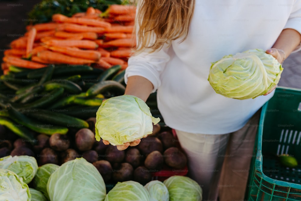 a woman holding two cabbages in front of a pile of vegetables