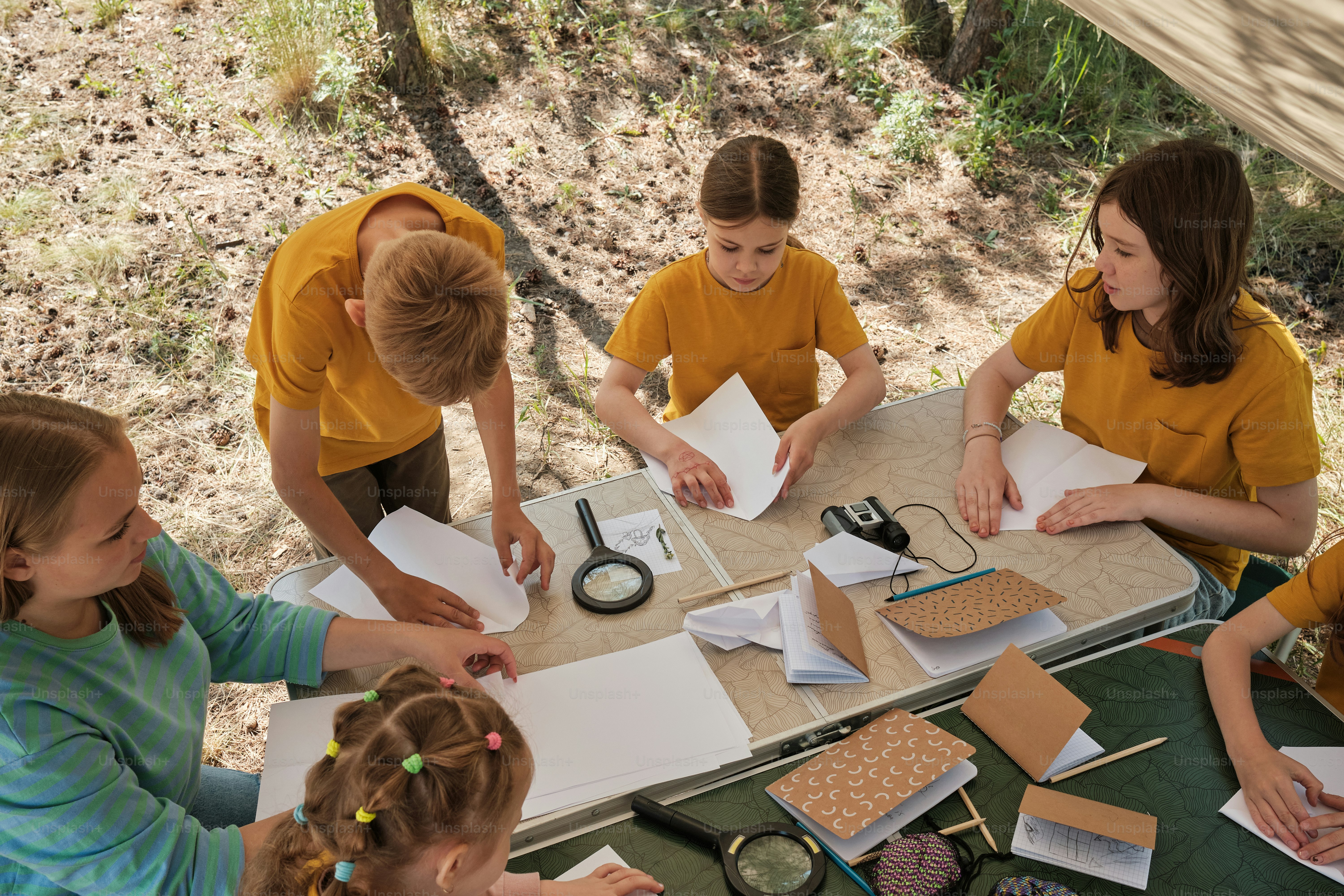 Children at summer camp making paper aeroplanes under a shelter in the forest.
