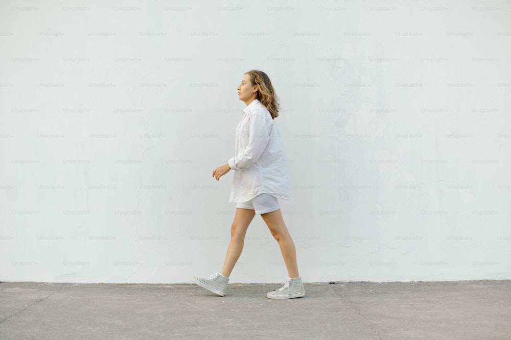 a woman walking down a sidewalk next to a white wall