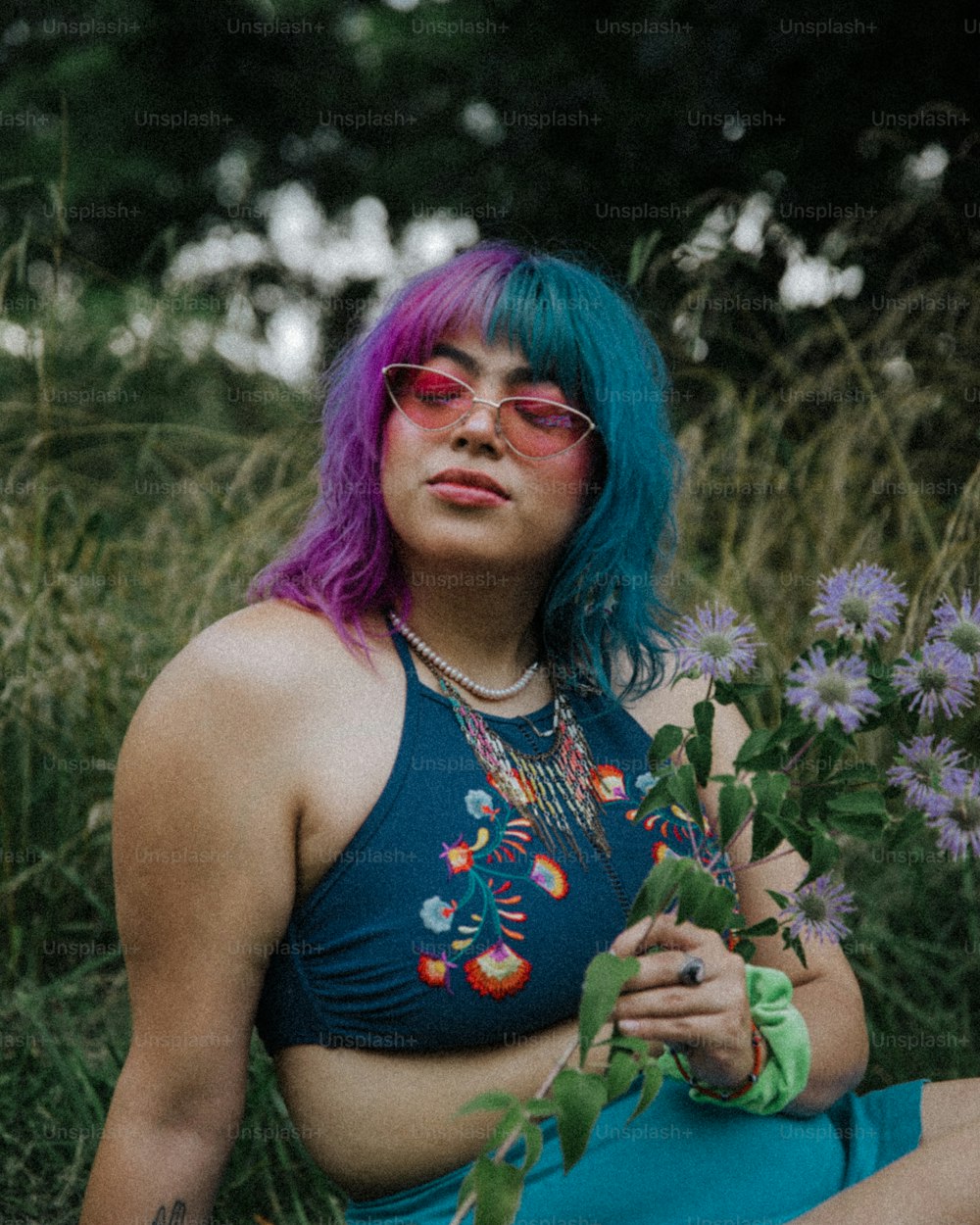 a woman with blue hair and pink glasses sitting in a field of flowers