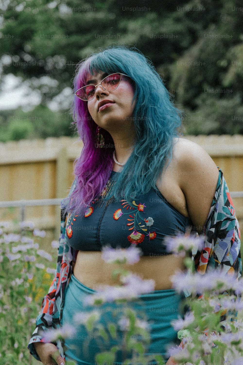 a woman with blue hair and glasses standing in a field of flowers