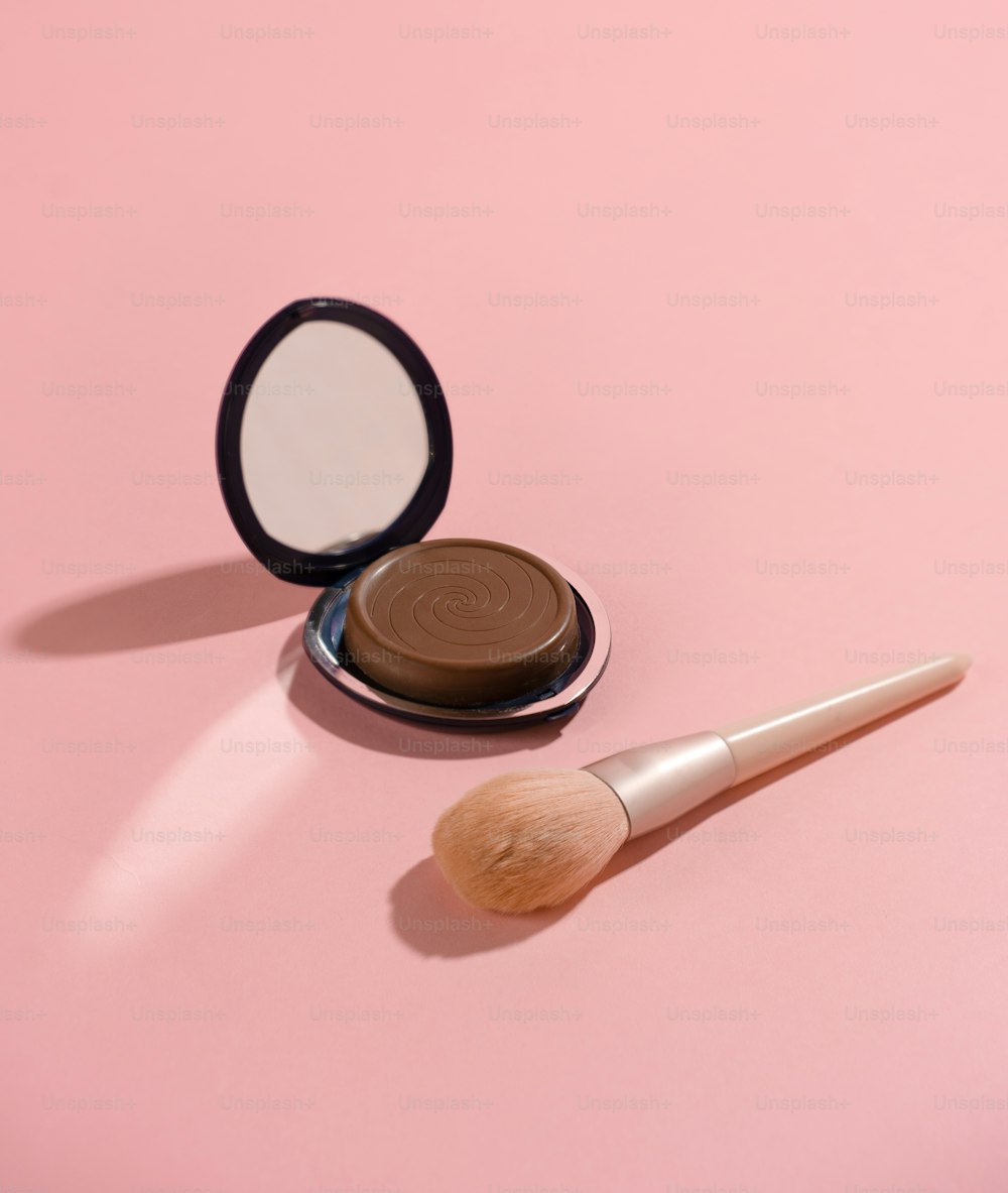 Un pincel de maquillaje sentado junto a un espejo compacto sobre una superficie rosada