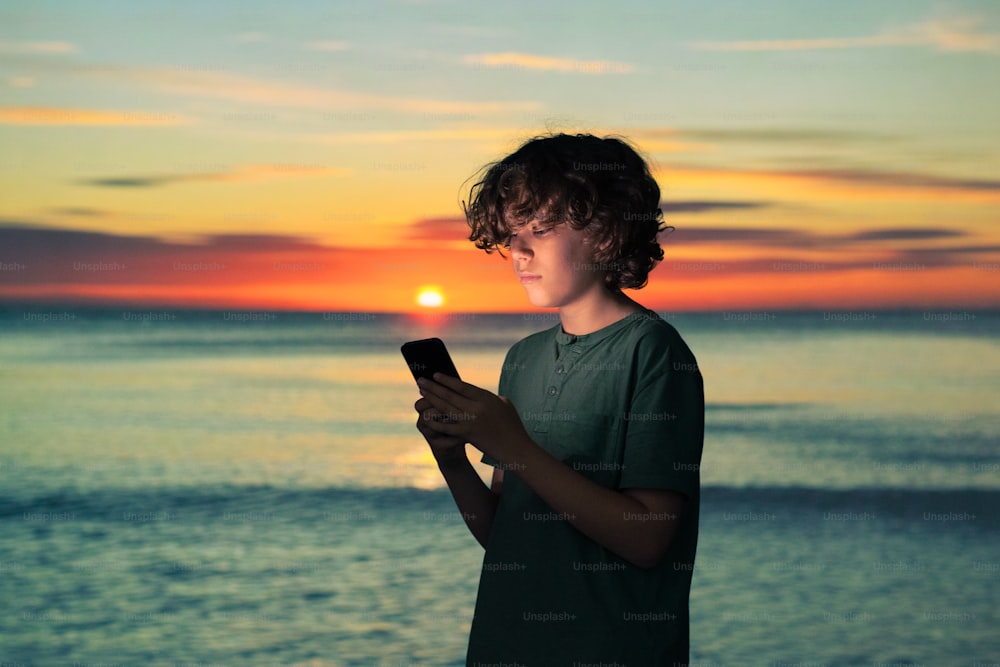 a young man standing on a beach holding a cell phone