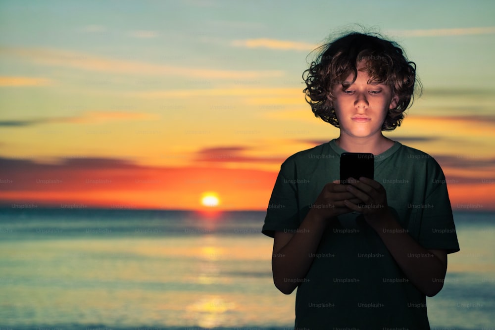a young boy holding a cell phone in front of a sunset