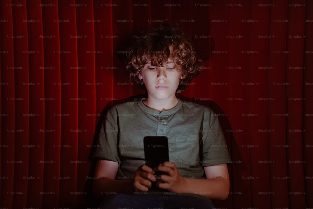 a young boy sitting in front of a red curtain holding a cell phone