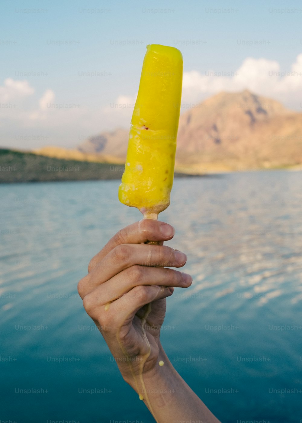 a hand holding a yellow popsicle over a body of water