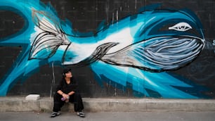 une personne assise sur un trottoir devant un mur avec un poisson peint sur