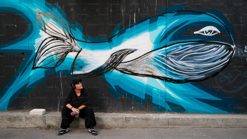 a person sitting on a curb in front of a wall with a fish painted on