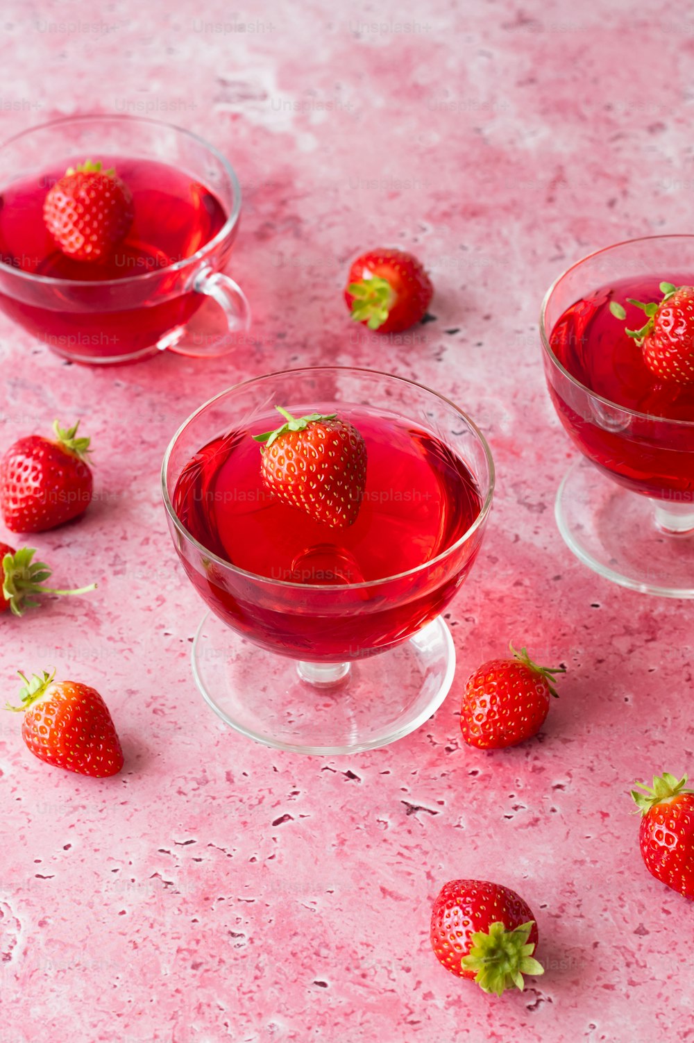 three glasses of red liquid with strawberries on a pink surface
