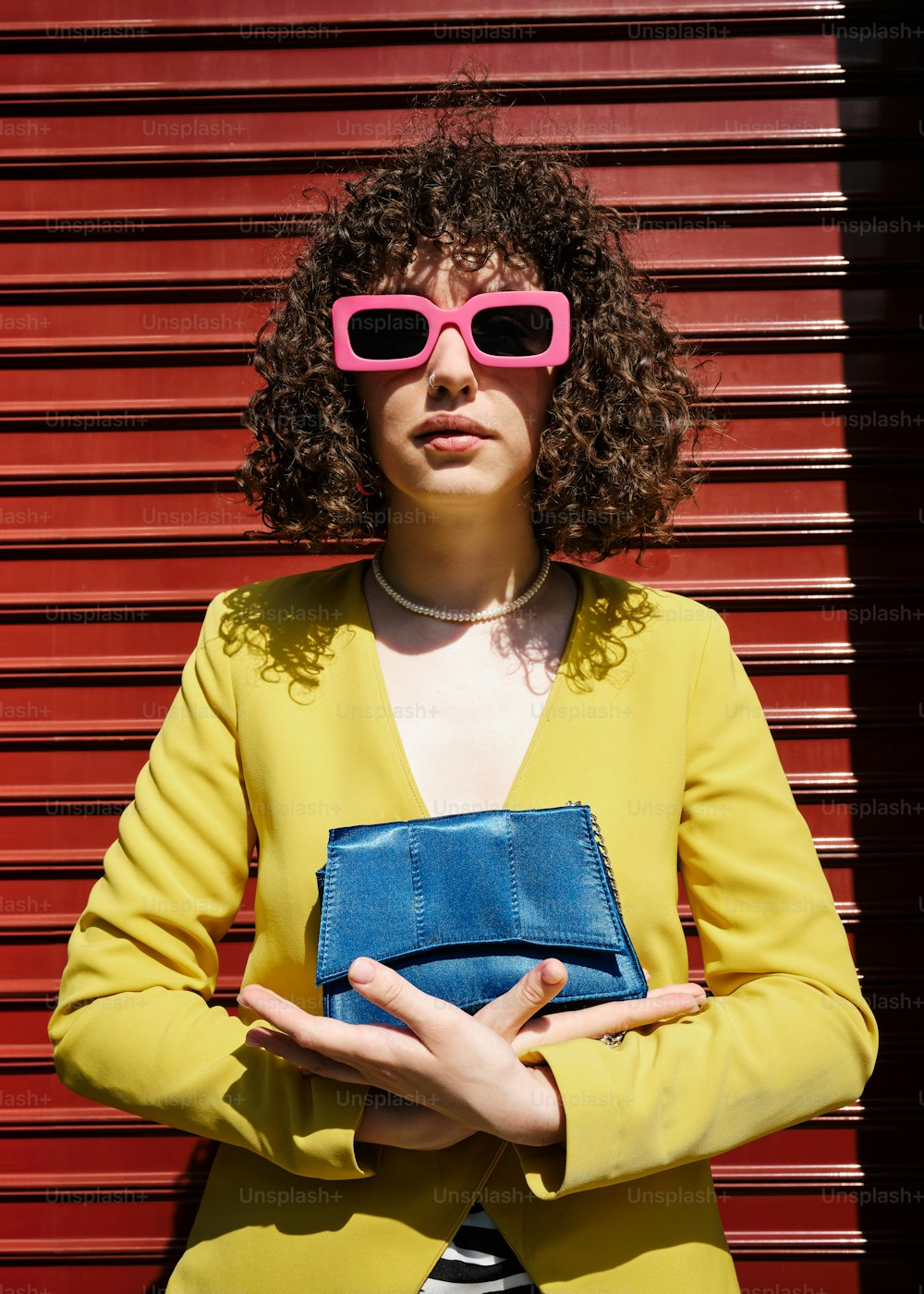 Una donna con una giacca gialla e occhiali da sole rosa che tiene una borsa blu