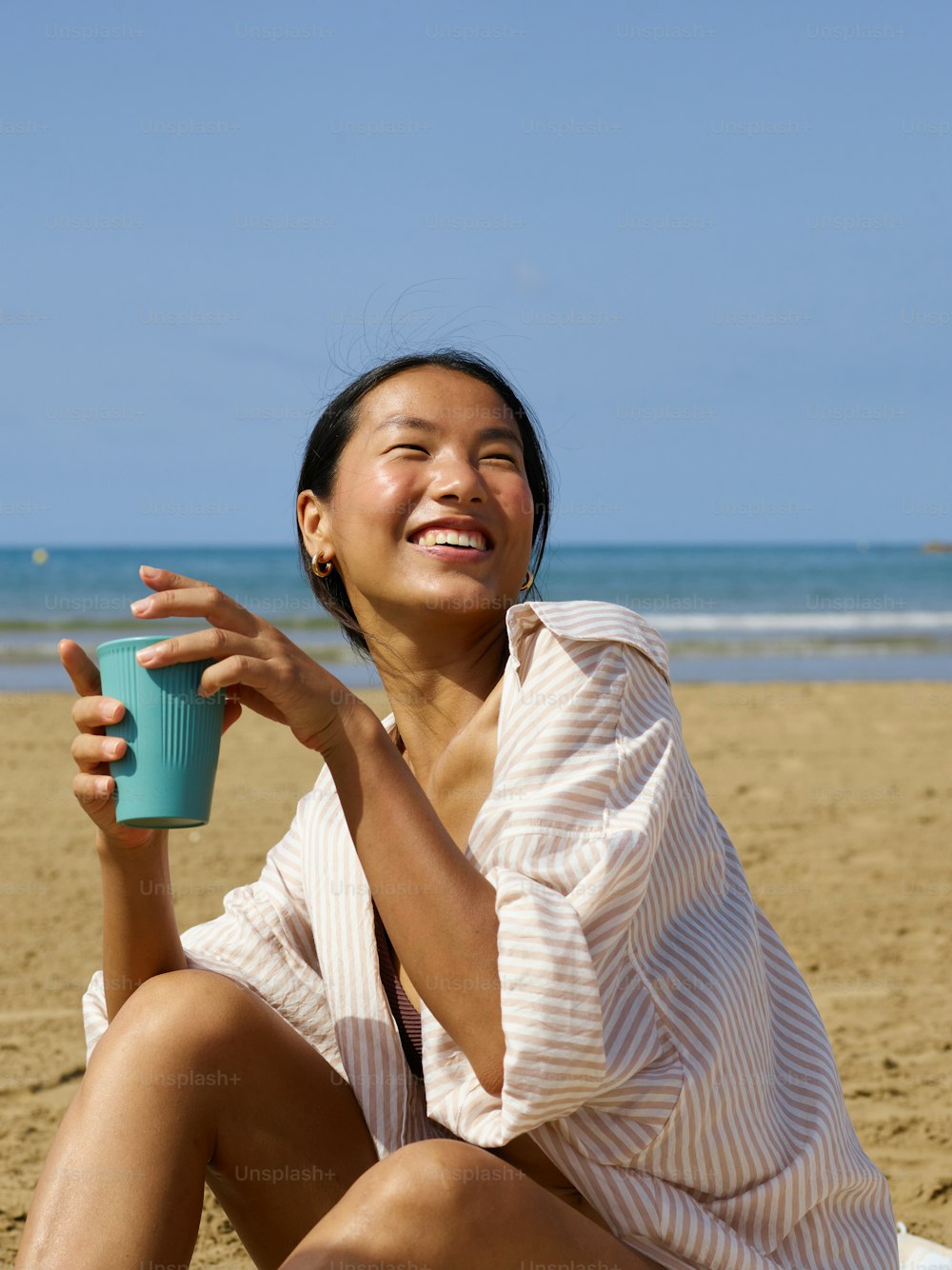 a woman sitting on the beach holding a cup of coffee