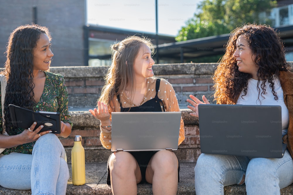 Tres mujeres jóvenes sentadas en escalones hablando y usando computadoras portátiles