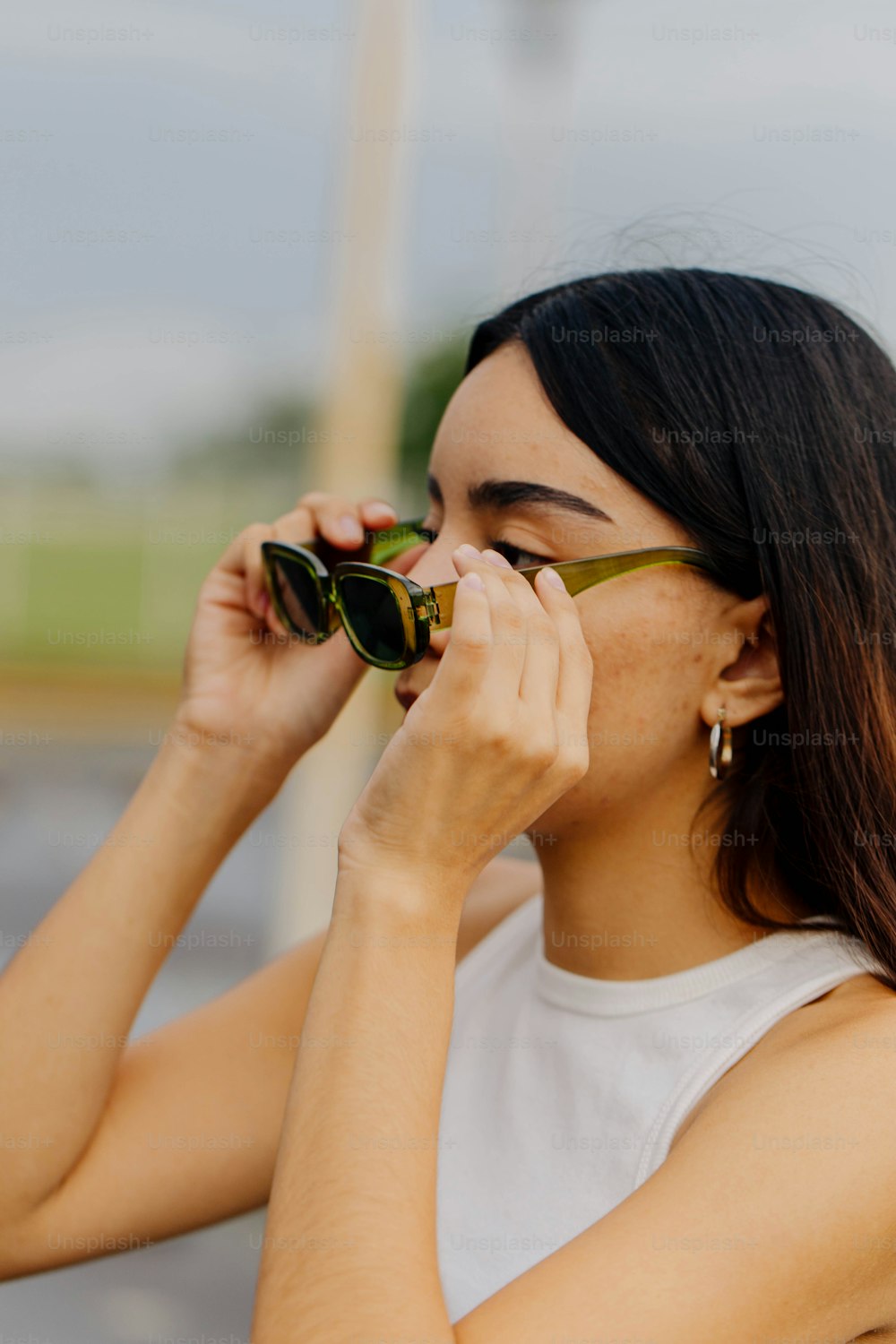 a woman in a white tank top is holding a pair of sunglasses to her face