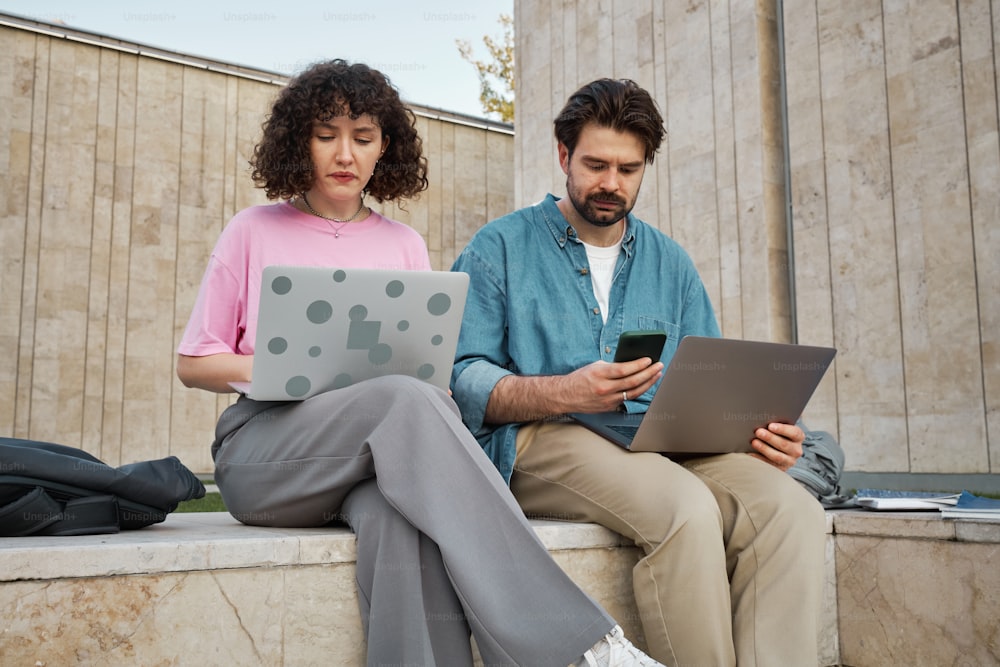 a man and a woman sitting on a bench looking at their laptops