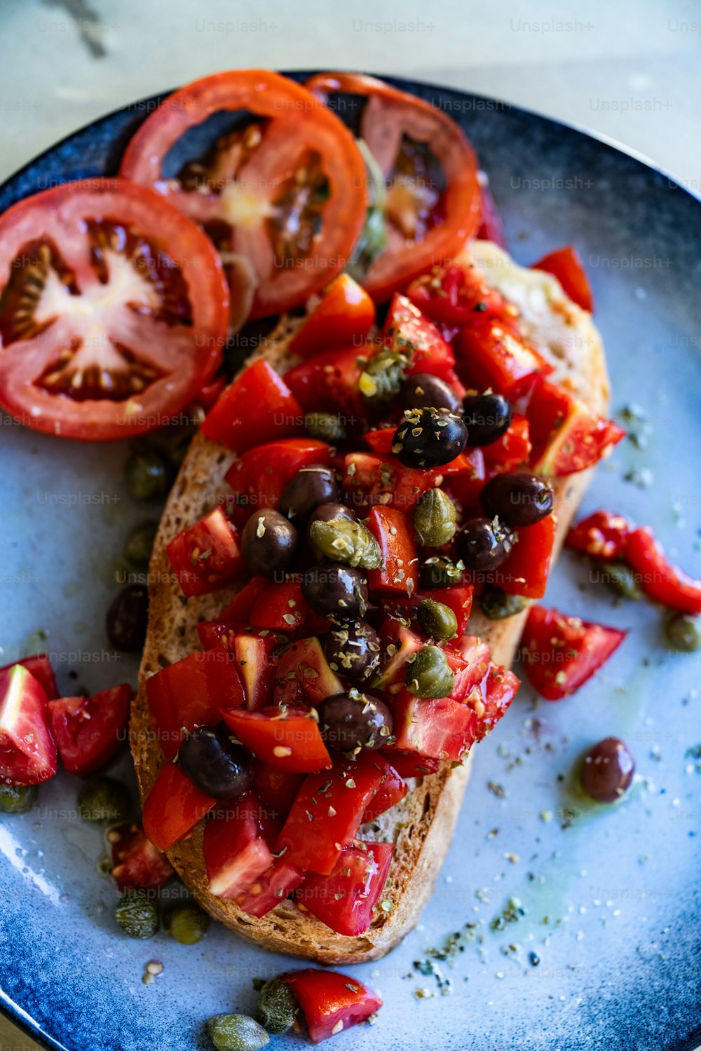 une assiette bleue surmontée d’un morceau de pain recouvert de tomates et d’olives