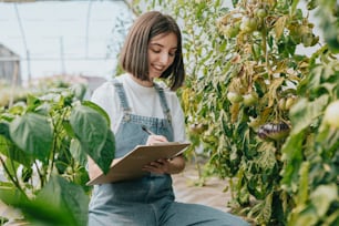 a woman in overalls writing on a clipboard in a greenhouse