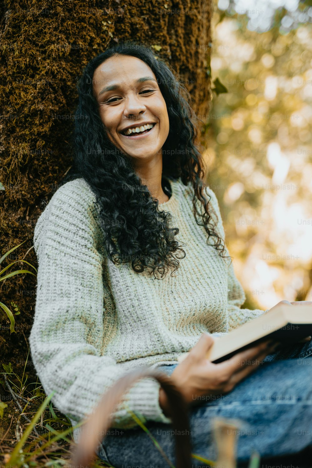 a smiling woman sitting on the ground next to a tree