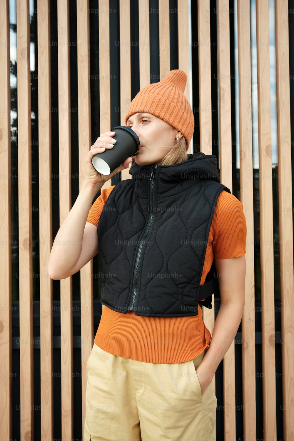 a woman drinking from a cup while standing in front of a wooden fence