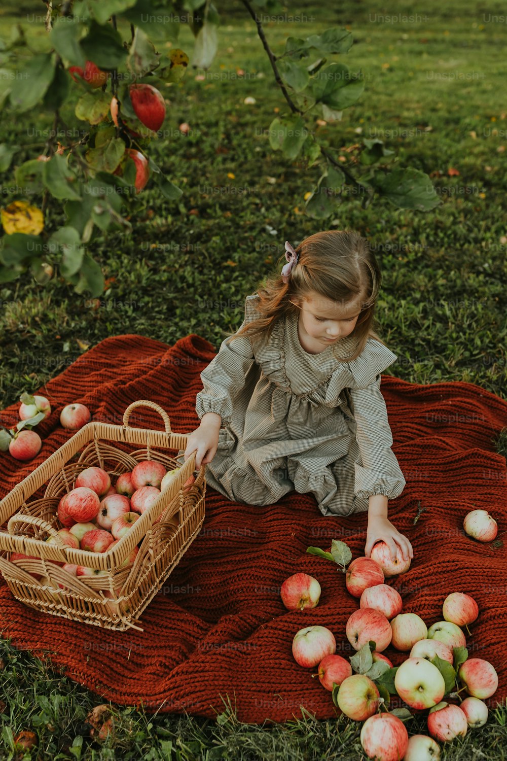a little girl sitting on a blanket with apples in a basket