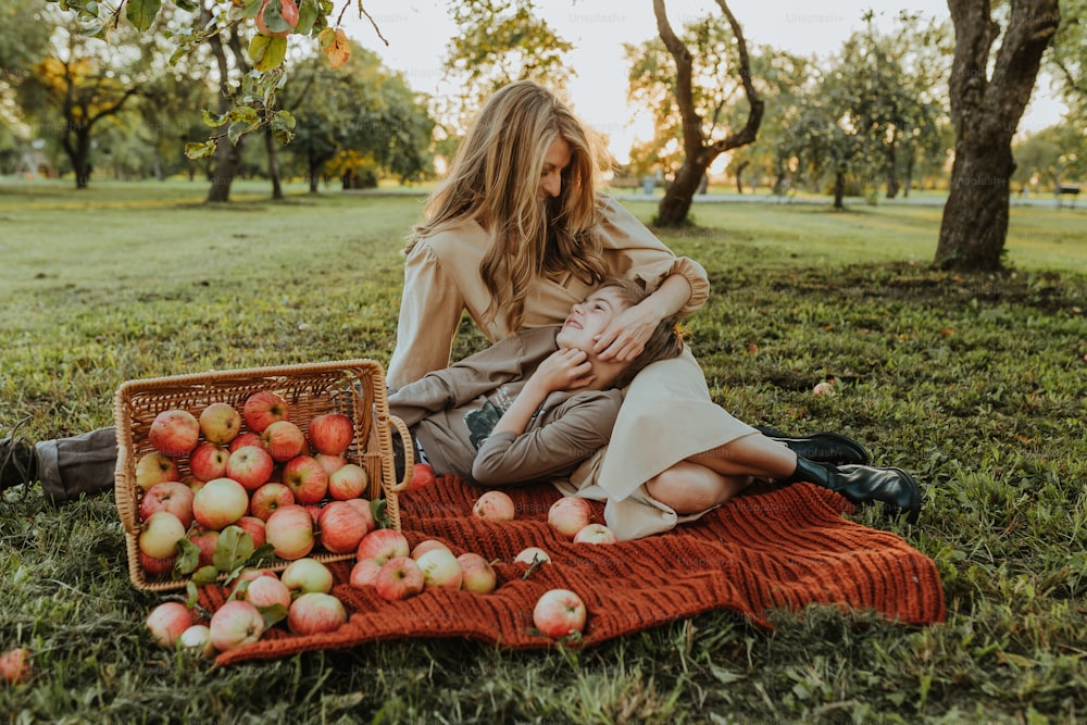 a woman holding a baby sitting on a blanket next to a basket of apples