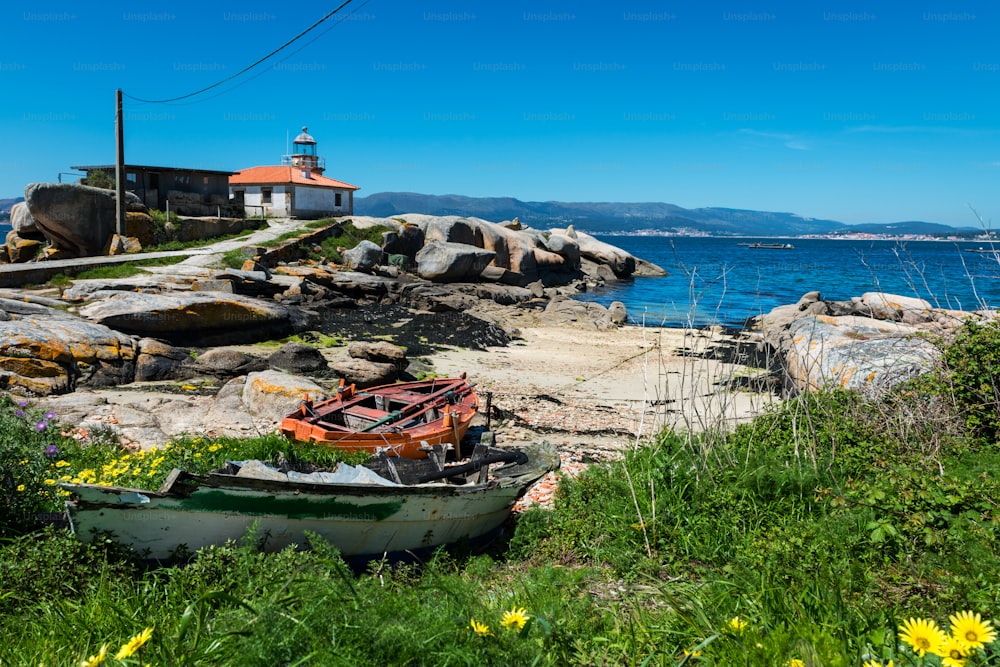 スペイン、ガリシアのリアスバイシャスにあるイラデアロウサ島にある小さな古い灯台で、前景にいくつかの放棄された木製のボートがあります。
