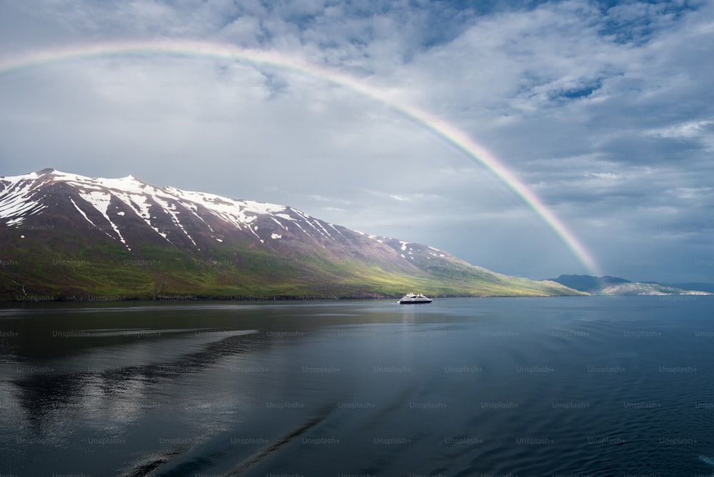 Ein atemberaubender Blick auf den Regenbogen über dem Meer in der Nähe der schneebedeckten Berge und ein einsames Schiff in Akureyri, Island
