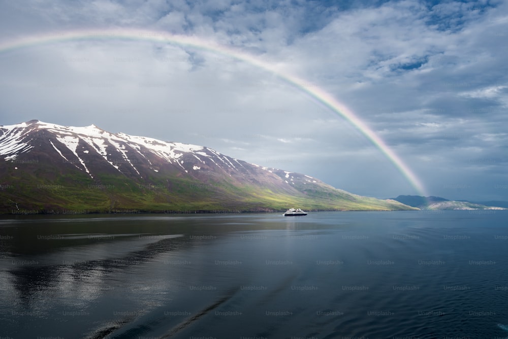 A breathtaking view of the rainbow over the sea near the snowy mountains and an isolated ship in Akureyri, Iceland