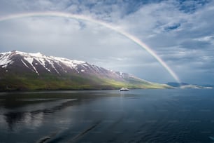 A breathtaking view of the rainbow over the sea near the snowy mountains and an isolated ship in Akureyri, Iceland