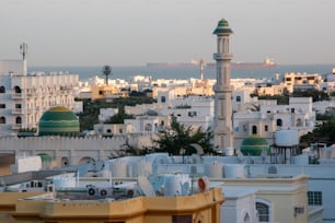 A beautiful shot of a white Mosque with green domes in the neighborhood with a sailing ships in the sea in the background on a sunny day