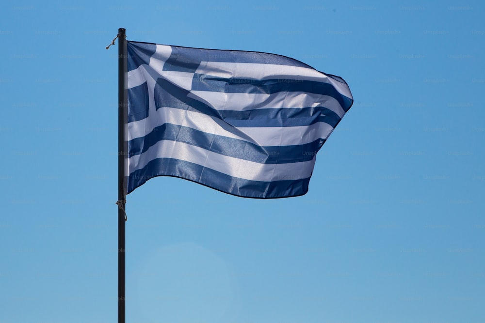 Uma visão de ângulo baixo na bandeira nacional da Grécia batendo ao vento em um mastro. Isolado contra um céu azul brilhante.