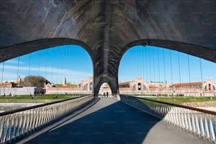 Lugareños y turistas caminan a través de un moderno puente sobre el río Manzanares en el parque público de Madrid Río en Madrid, España, con el edificio Matadero (un antiguo matadero convertido en un centro de arte) al fondo. 
